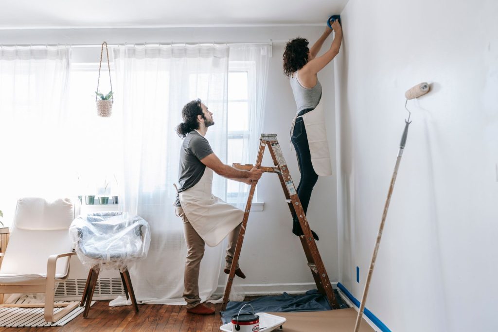 Couple managing some non-structural home repairs from their cash-out refinance. The woman is on the construction ladder putting a painter's tape on the wall while her husband is holding the ladder
