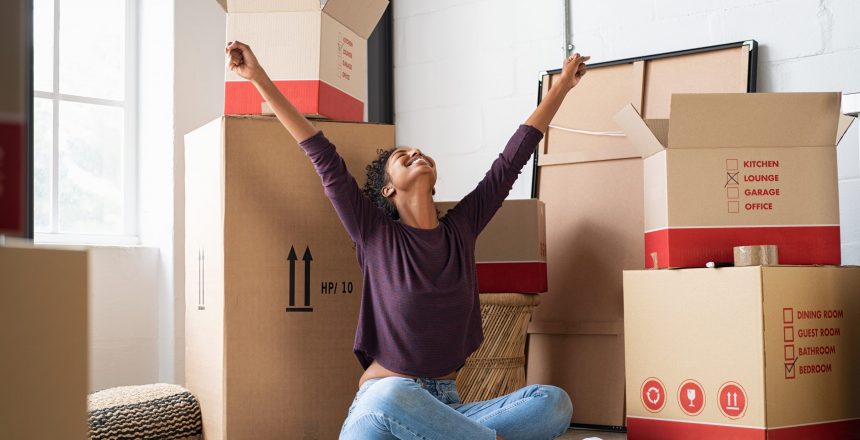 Happy young woman sitting in new apartment and raising arms in joy after moving in. Joyful and excited african girl moving to new home. Black woman sitting on floor in her house.